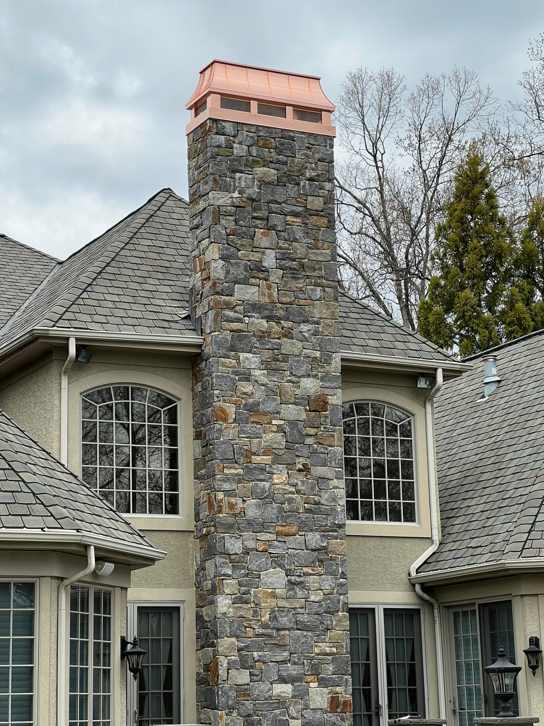 Custom copper chimney cap installed on tall stone masonry chimney — hand-fabricated by Archaic Metal for contractor direct order