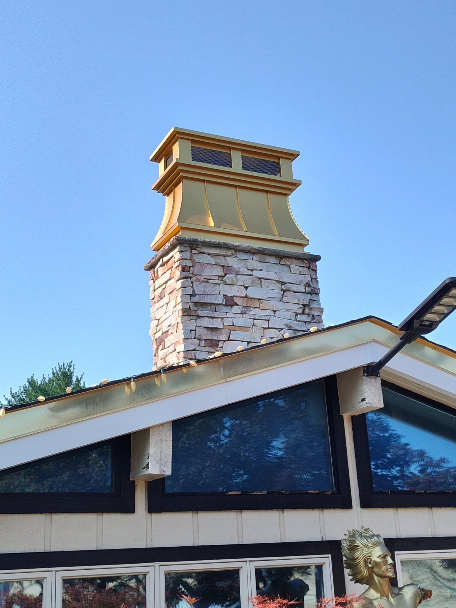 Close-up of gold anodized aluminum curved panel chimney cap on stone chimney — standing seam detail and crown molding visible, custom fabricated by Archaic Metal