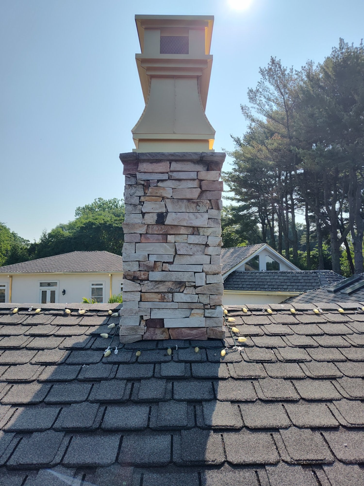 Gold anodized aluminum chimney cap installed on tall stone chimney, angled rooftop view — custom sized curved panel cap by Archaic Metal