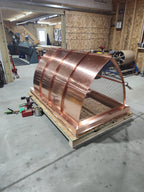 Large custom curved panel chimney cap in 16oz copper on a shipping crate — front angle view showing concave mansard profile and stainless steel spark screen, fabricated in the Archaic Metal shop in Berlin, NJ