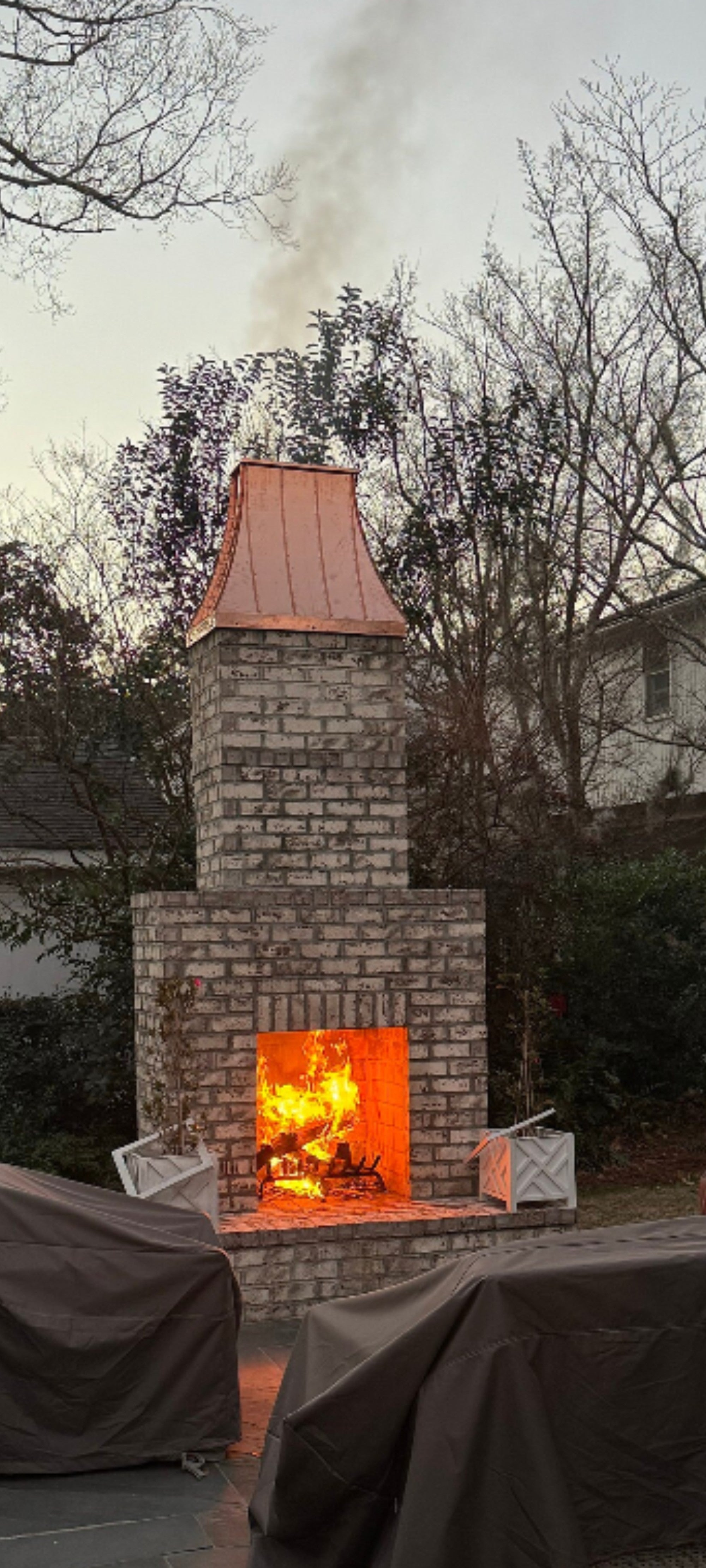  Custom copper chimney shroud on outdoor brick fireplace at dusk with fire burning, hand-fabricated by Archaic Metal