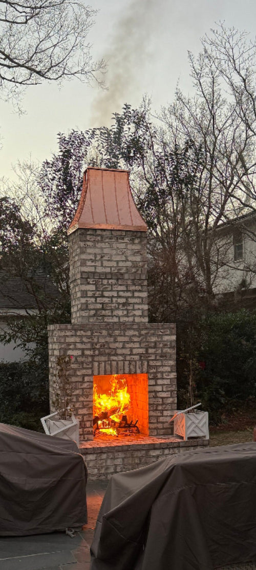  Custom copper chimney shroud on outdoor brick fireplace at dusk with fire burning, hand-fabricated by Archaic Metal