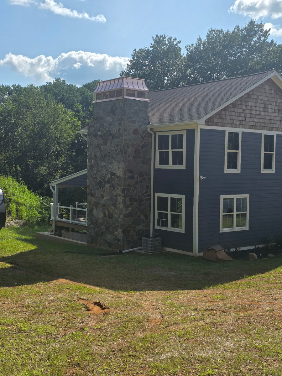 Custom copper chimney cap on tall stone chimney of blue shingle-sided home — hand-fabricated by Archaic Metal