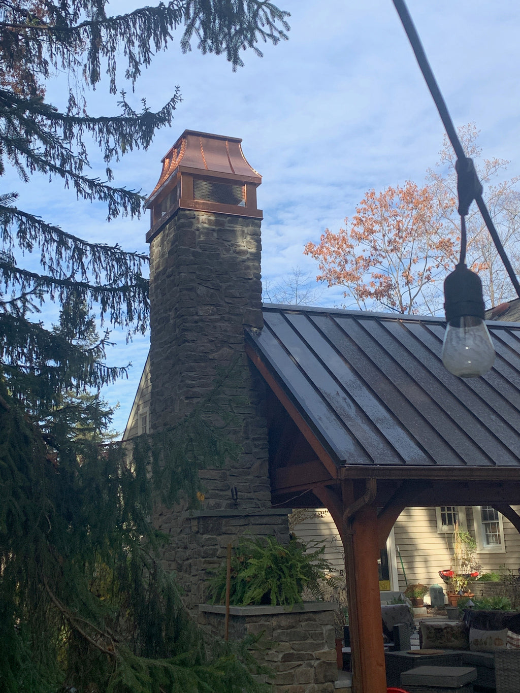 Custom copper chimney cap on tall stone chimney above outdoor timber pavilion with standing seam roof — hand-fabricated by Archaic Metal