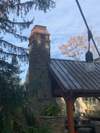 Custom copper chimney cap on tall stone chimney above outdoor timber pavilion with standing seam roof — hand-fabricated by Archaic Metal