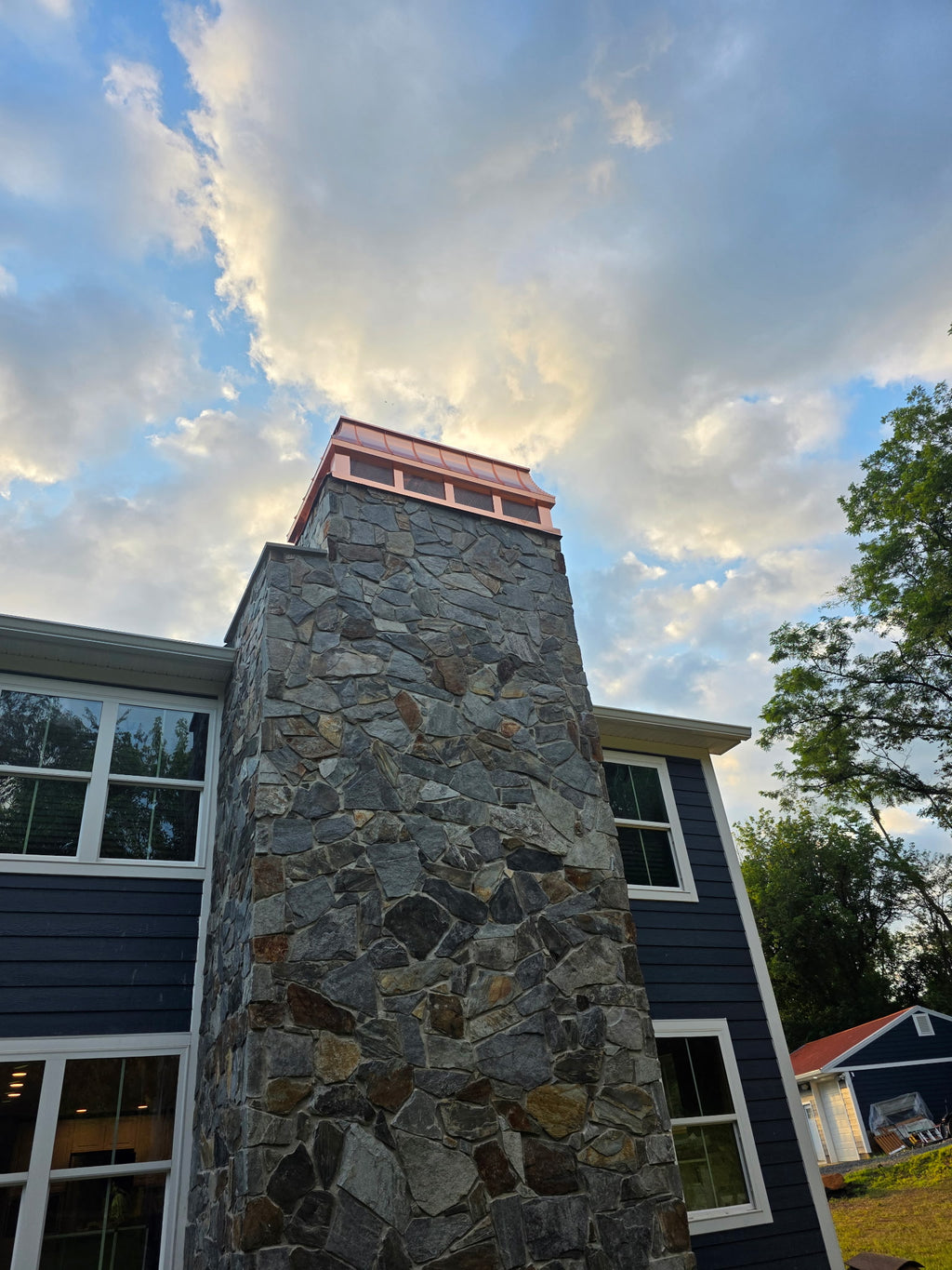 Custom copper chimney cap atop tall stone chimney on blue shingle home — dramatic sky, ground-level view, hand-fabricated by Archaic Metal