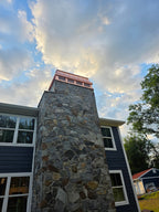 Custom copper chimney cap atop tall stone chimney on blue shingle home — dramatic sky, ground-level view, hand-fabricated by Archaic Metal
