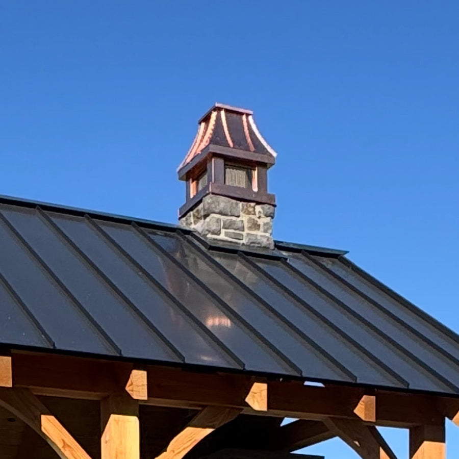 Custom copper curved panel chimney cap on stone chimney above timber frame outdoor pavilion with standing seam roof — hand-fabricated by Archaic Metal