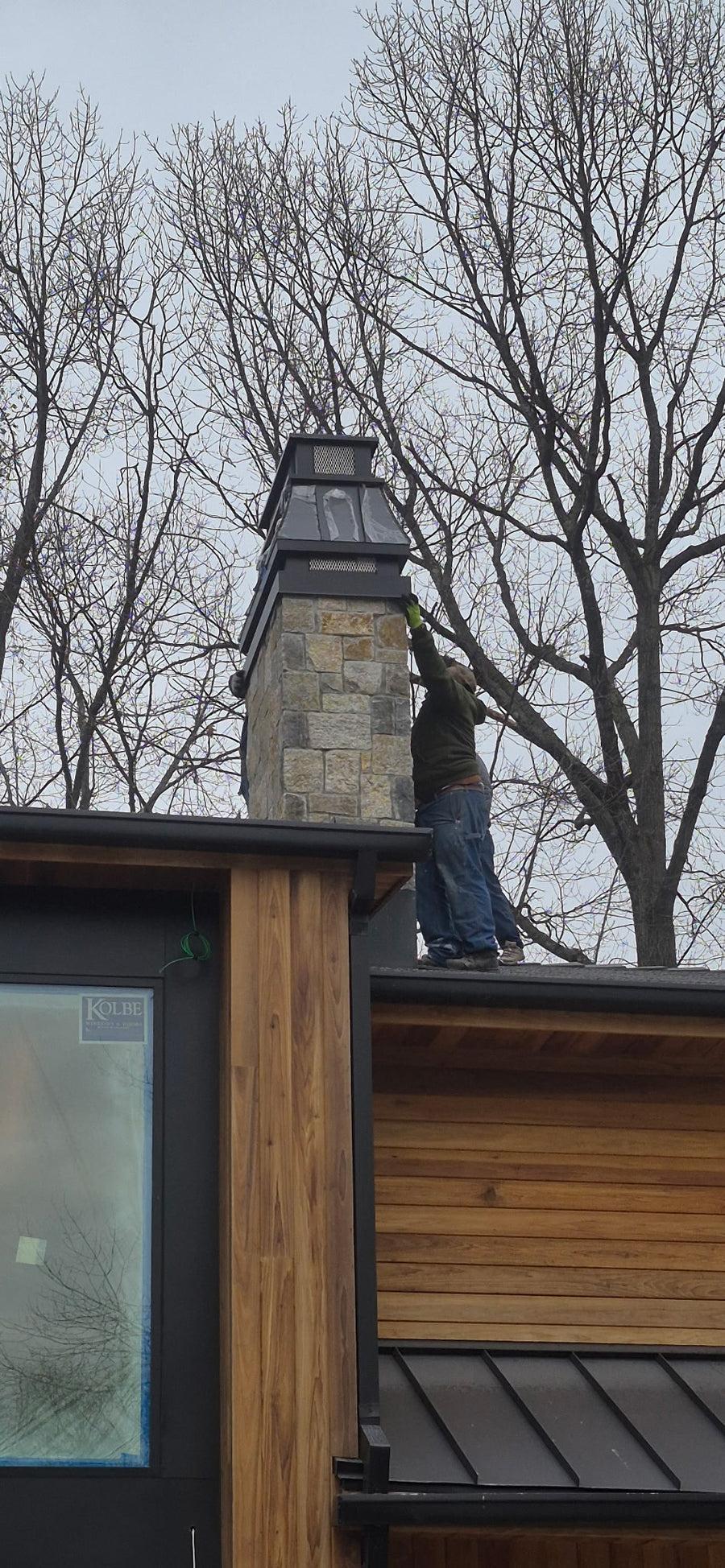 Contractor installing custom Kynar black chimney cap on stone chimney of modern wood and steel home — hand-fabricated by Archaic Metal