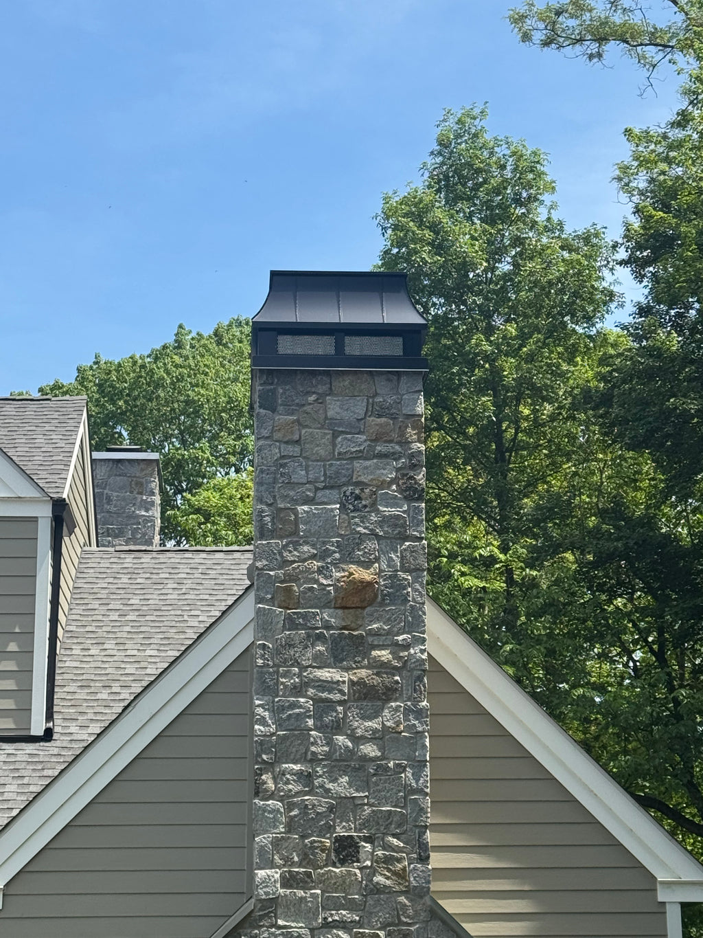 Custom Kynar black straight panel chimney cap with spark screen on stone masonry chimney above shingle roof — hand-fabricated by Archaic Metal