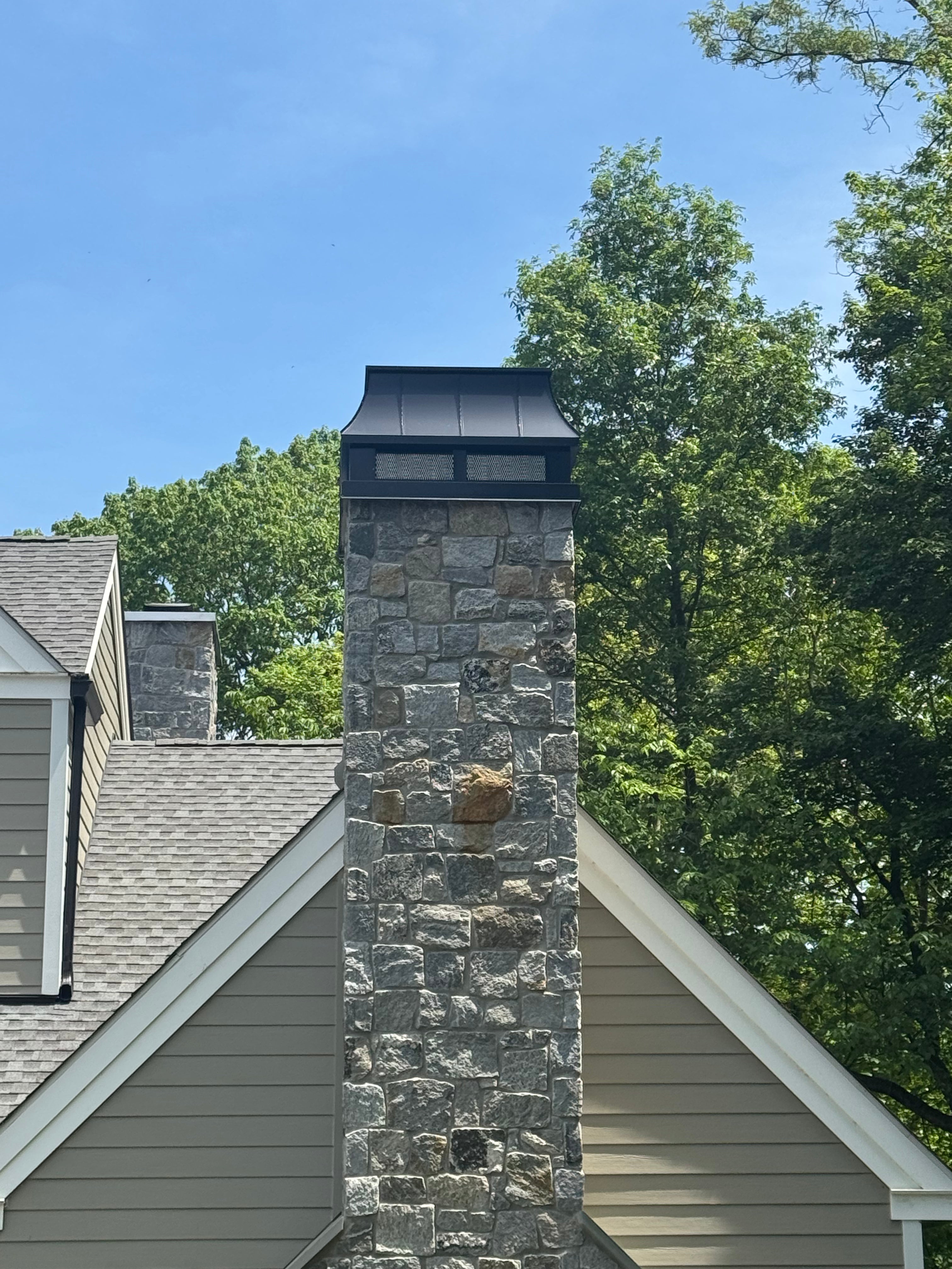 Custom Kynar black straight panel chimney cap with spark screen on stone masonry chimney above shingle roof — hand-fabricated by Archaic Metal