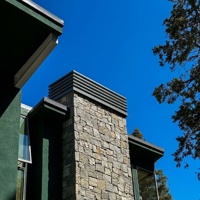 Louvered chimney cap installed on stone chimney against blue sky — custom fabricated by Archaic Metal